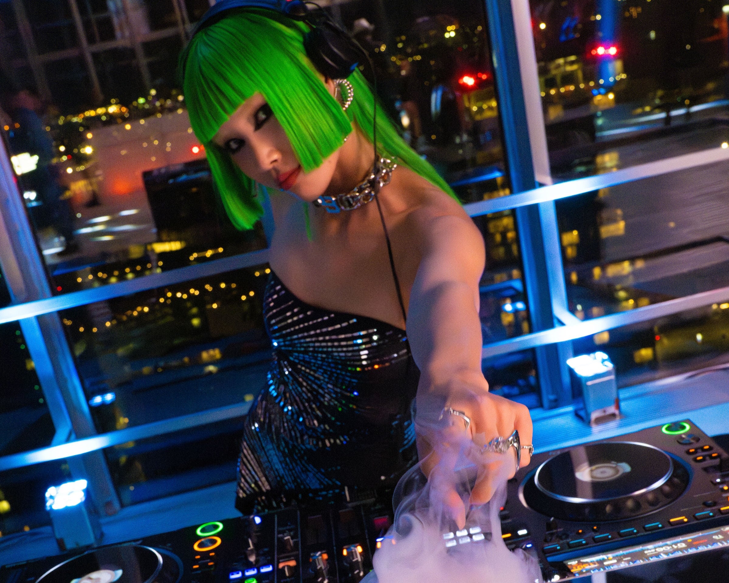 Person with green hair at a DJ booth with cityscape in the background