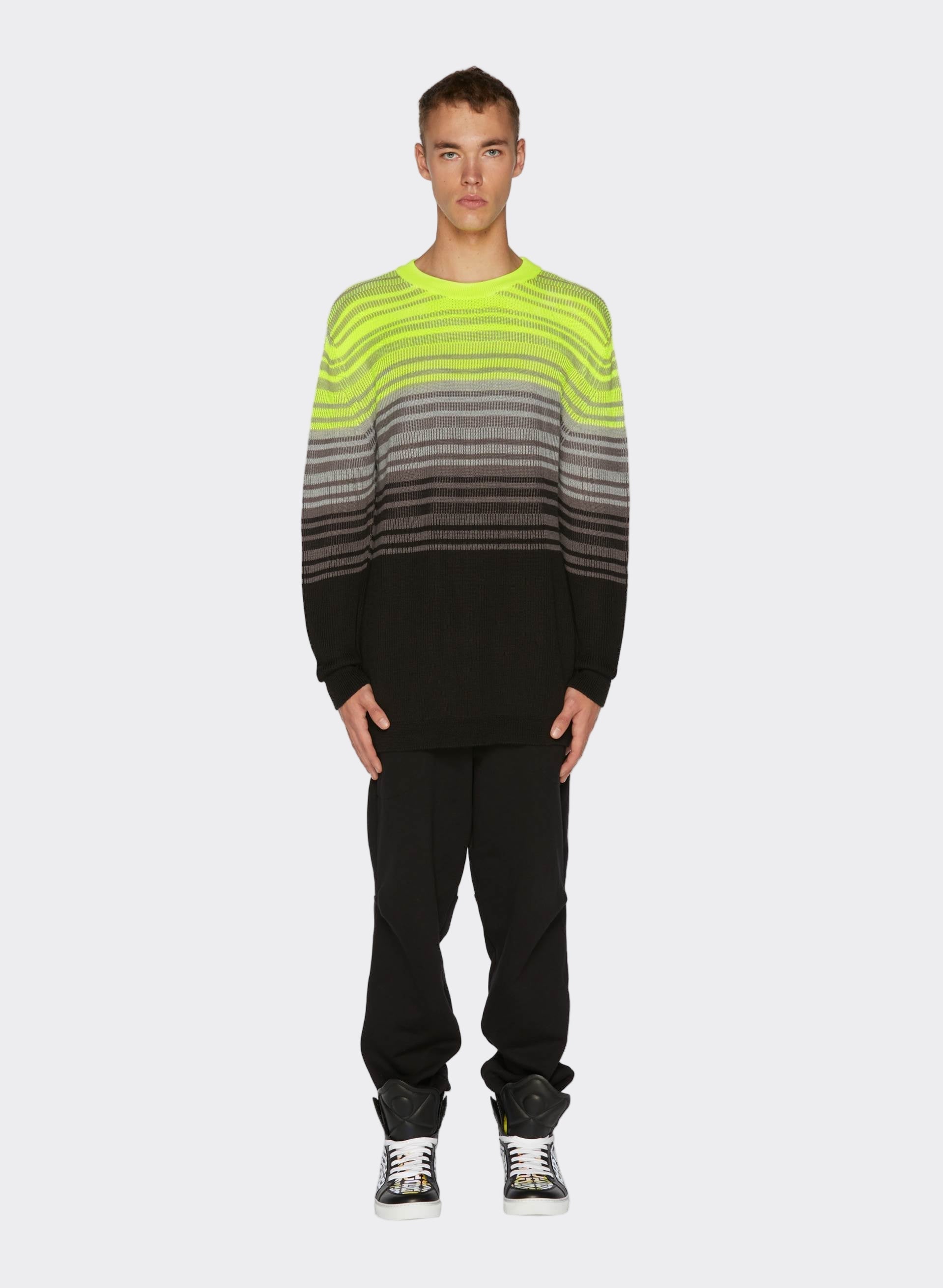 Model wearing a Knit Fluo Crewneck in neon green, gray, and black, paired with relaxed black trousers and 3D Logo Iridescent Sneakers
