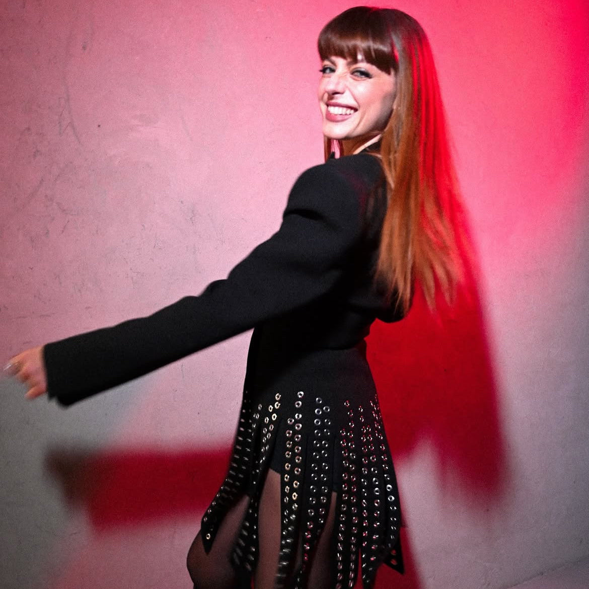 Woman in a black outfit with fringe and embellishments against a pink wall.