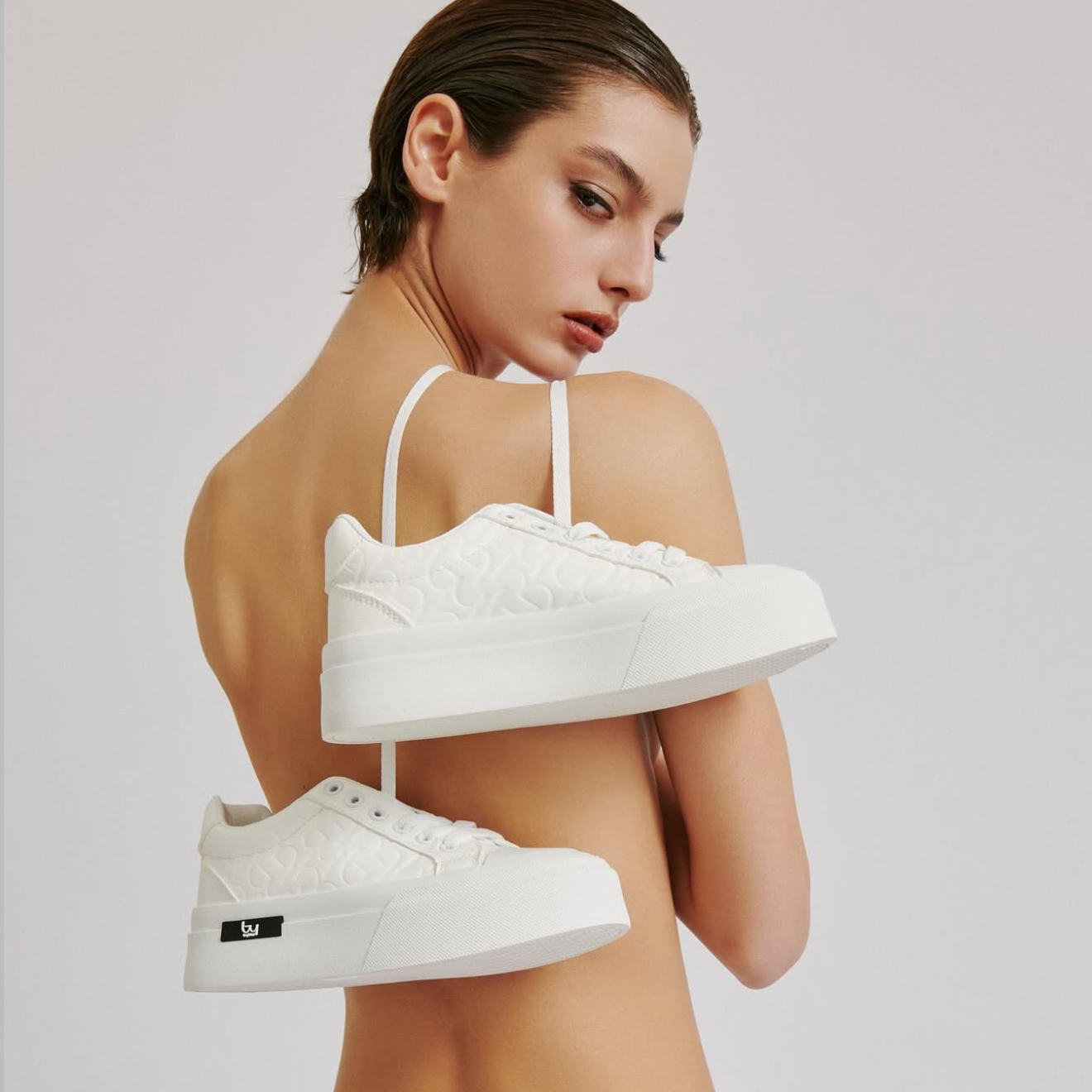 Person wearing a white bra and holding white sneakers on a plain background
