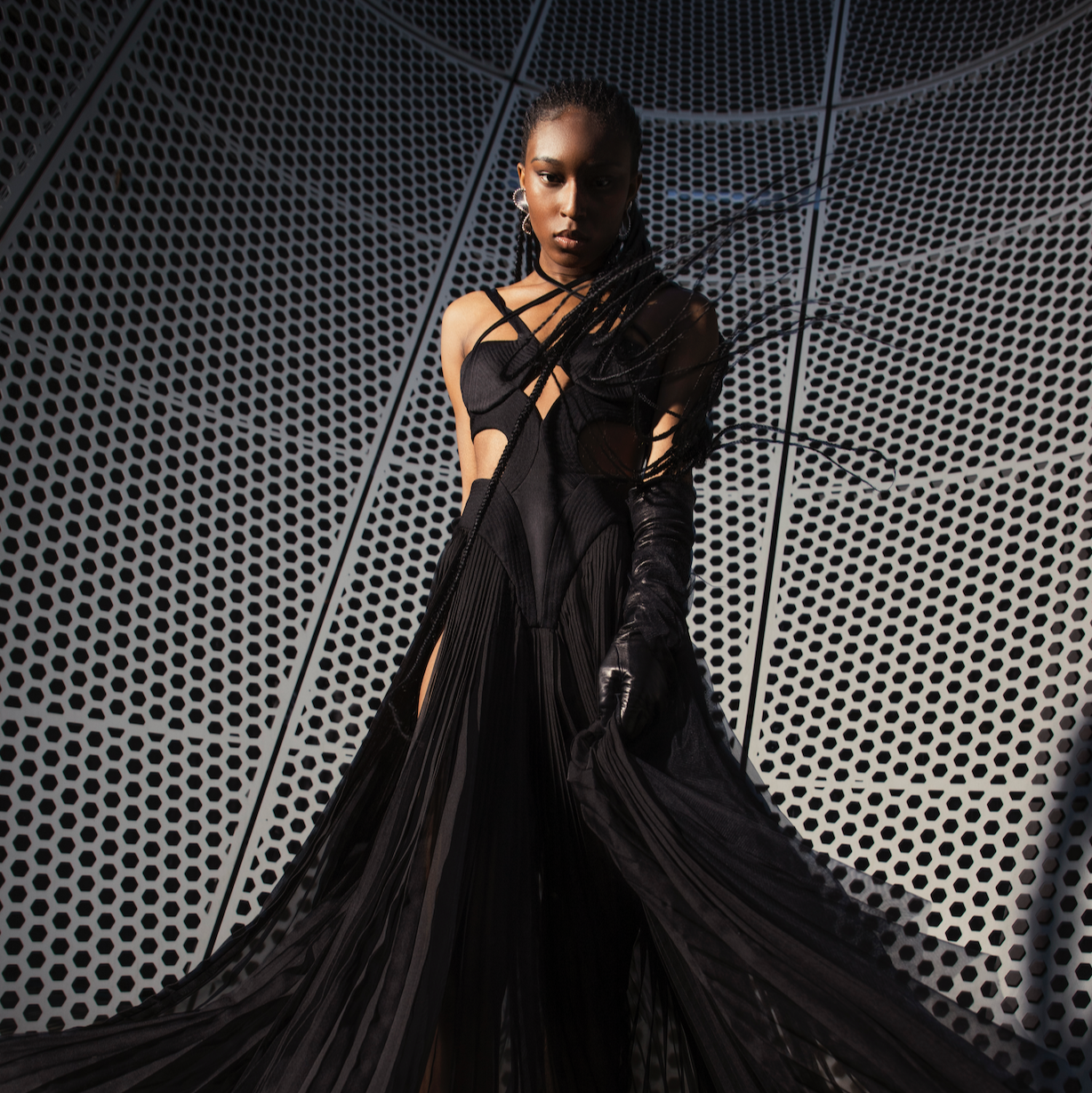 Model wearing a long black dress with intricate detailing against a geometric patterned wall.