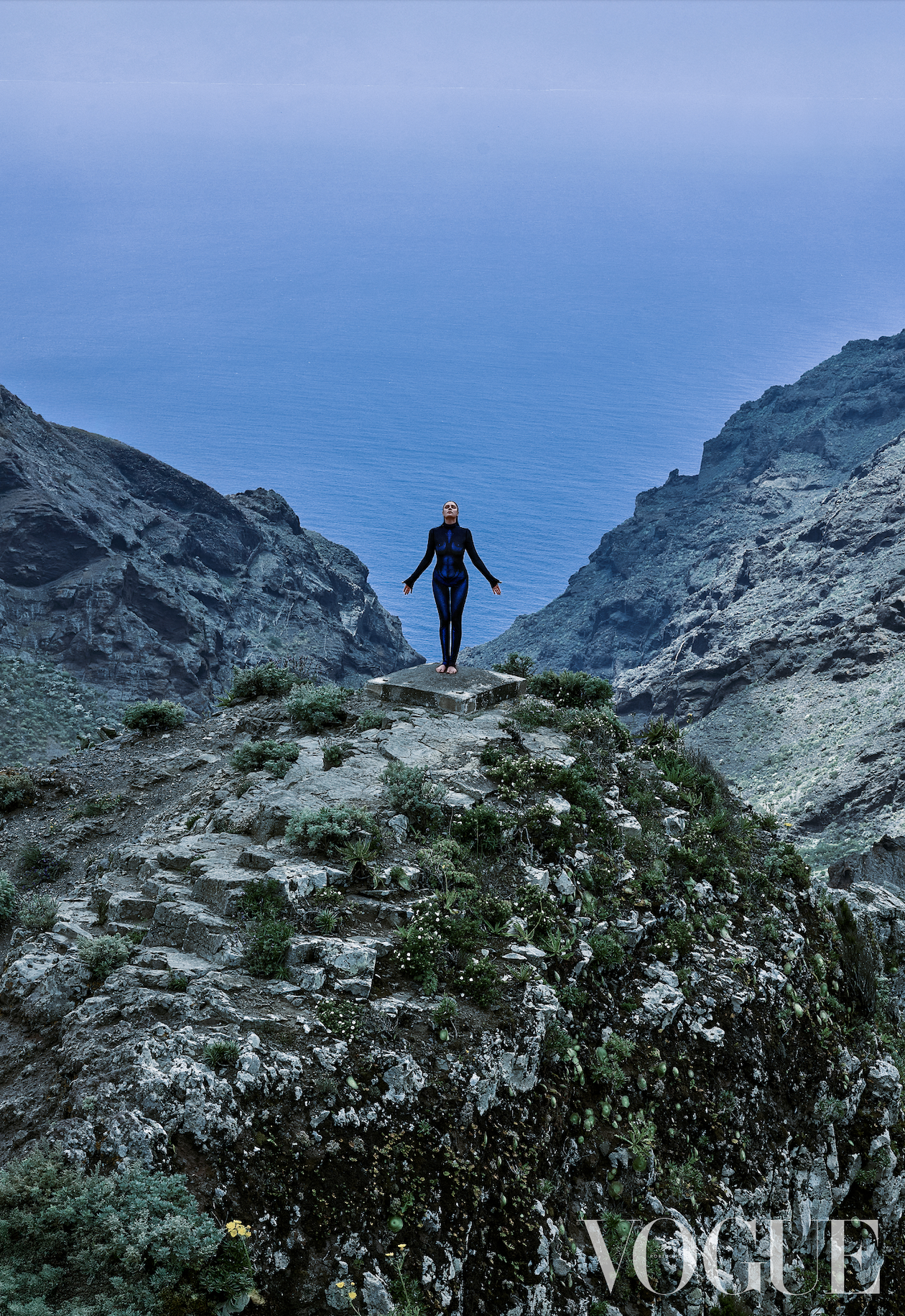 Person standing on a rocky outcrop with mountains in the background, Vogue logo visible